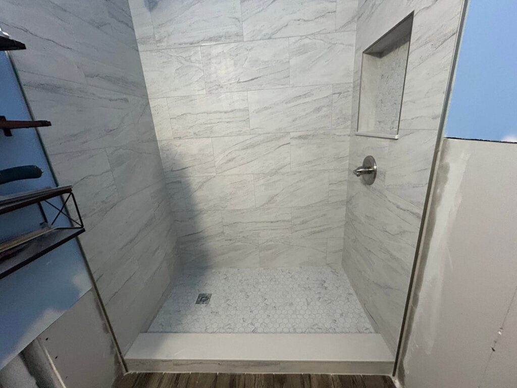 Shower with light gray tile walls, built-in shelf, metal faucet, and unfinished walls outside the shower.