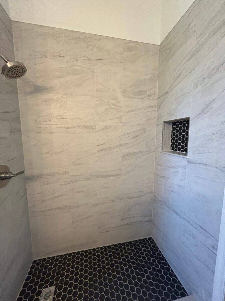 Shower with light gray rectangular tiles, black hexagonal floor tiles, and a small recessed shelf with black hex tile.