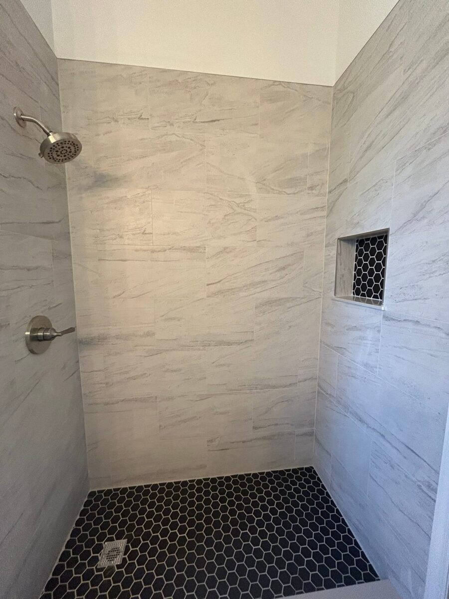Shower with light gray wall tiles, black hexagon floor tiles, a silver showerhead, and a built-in wall niche.
