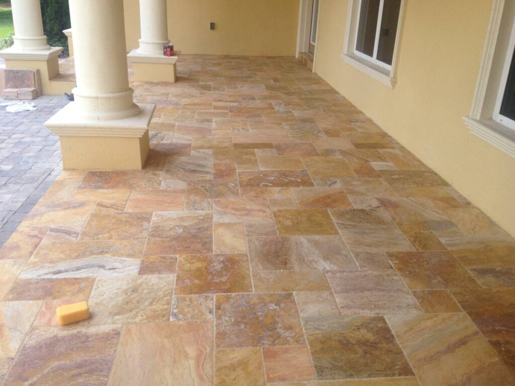 Tan and beige stone tiles cover a patio area next to columns and windows, with a sponge on the floor.